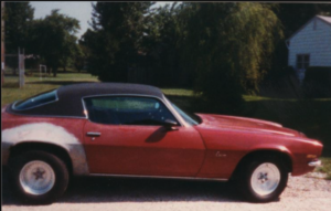 Sideshot of 1970 Camaro. It is red with a black vinyl top, with lots of bondo around the rear wheels. It has big tires in the back and skinny tires in the front.