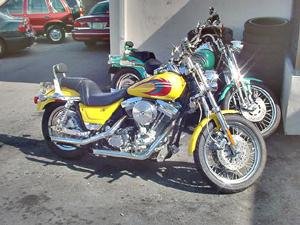 Two motorcycles parked side by side. The closer motorcycle is a yellow Harley FXR4